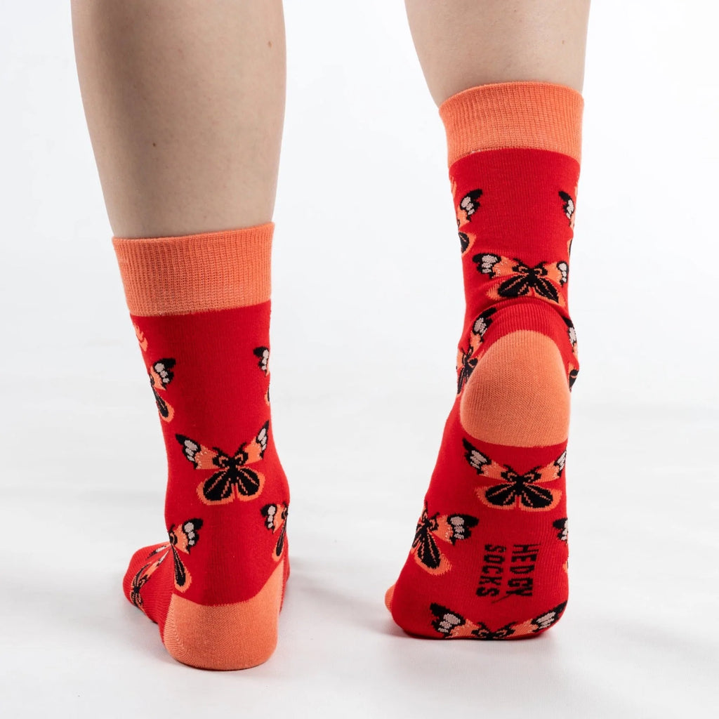 Back end view of model walking away in super soft premium quality bamboo socks with an orange and red butterfly print on an orange background and brand logo on sole of foot